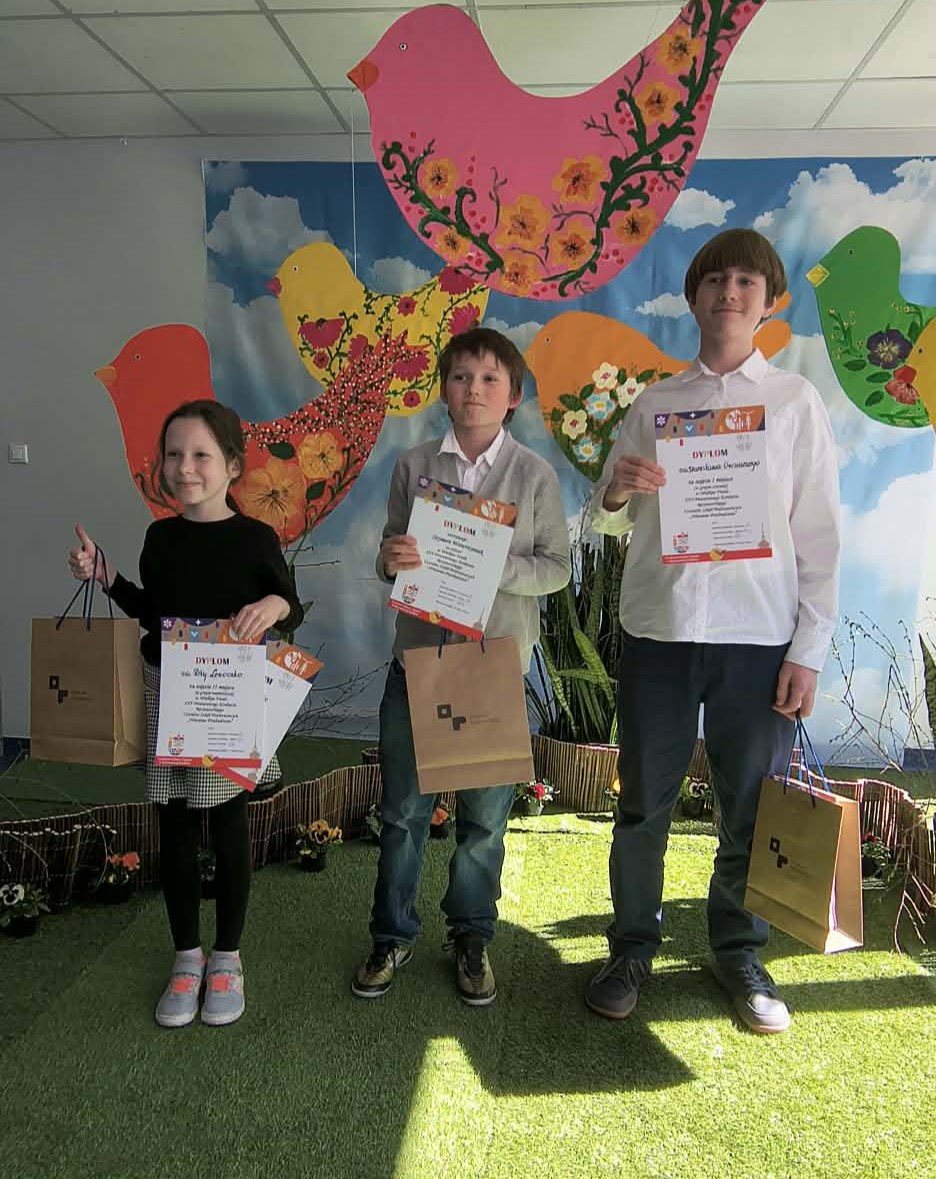 Przedstawia tr&oacute;jkę dzieci z dyplomami na tle wiosennej scenografii